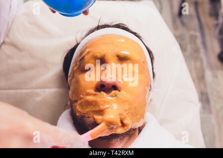 Die Kosmetikerin stellt eine kosmetische Maske auf das Gesicht des Client mit Hilfe einer medizinischen Spachtel männlich Mann. Stockfoto