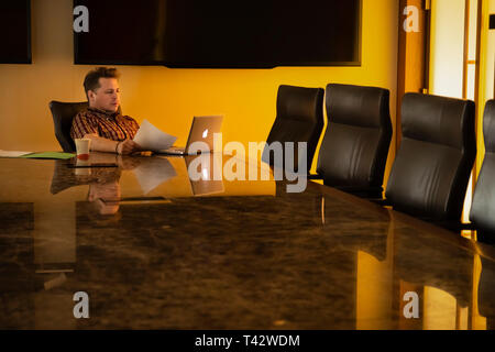 Newark, NJ - 12. April 2019: der Mensch arbeitet am Laptop zu großen Konferenztisch; am späten Abend oder frühen Morgen scheint die Sonne durch das Fenster. Stockfoto