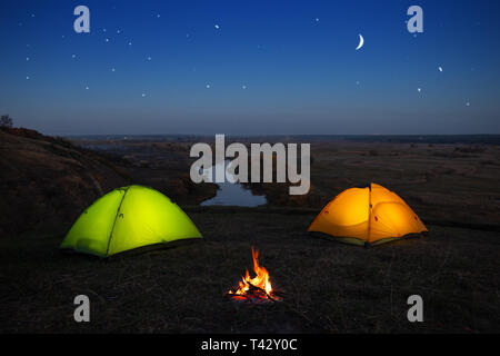 Orange und grüne Zelt durch den Fluss in der Nacht. Camping von zwei zelten und Lagerfeuer am Ufer unter einem Sternenhimmel. Sommer Landschaft. Konzept der Tr Stockfoto