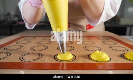 Konditor gießt Teig aus gebäckbeutel auf Schablone Silikon Matte auf dem Tisch. Kochen Makronen. Die Hände schließen. Stockfoto