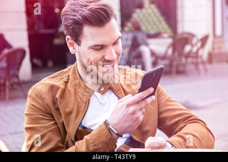 Glückliche junge Erwachsene charmante Mann auf dem Smartphone auf öffentlichen Café Terrasse Stockfoto
