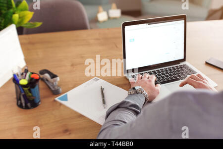 Mann bei der Arbeit auf seinem Laptop im Büro, verschwommenes Notepad, Markierungen und Kalender auf dem Schreibtisch, Ansicht von hinten, Detail und Armbanduhr Stockfoto