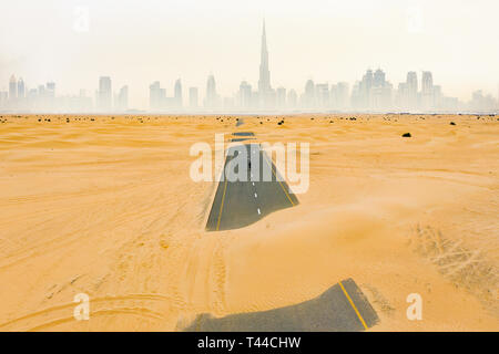 Beeindruckende Luftaufnahme von einer nicht identifizierten Person zu Fuß auf einer verlassenen Straße durch die Sanddünen in Dubai Wüste bedeckt. Skyline von Dubai durch Nebel umgeben Stockfoto