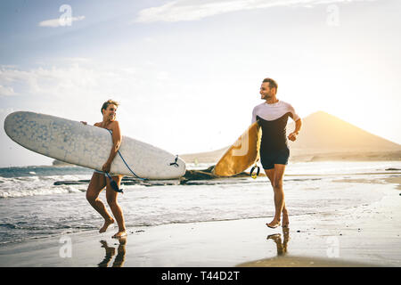 Junge paar Surfer mit surfbrettern am Strand bei Sonnenuntergang - glückliche Liebhaber zu surfen gehen zusammen - Menschen, Sport und Lifestyle Konzept