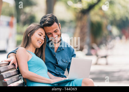 Glückliches junges Paar mit Laptop sitzen auf einer Bank in der Stadt im Freien - Zwei Liebende Spaß Zeit miteinander zu verbringen, soziale Netzwerk zu beobachten Stockfoto