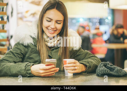 Junge schöne Frau trinkt einen Kaffee in einem Café Bar während der Eingabe auf mobilen Smart Phone Application Chat - hübsches Mädchen mit einem heißen Getränk Stockfoto