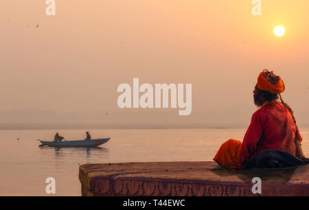 Hindu sadhu Baba in Meditation am Ganges River Bank bei Sonnenaufgang mit Blick auf einen hölzernen Boot auf dem Fluss Ganges Stockfoto