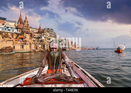 Sadhu Baba in Meditationshaltung auf einer hölzernen Boot auf dem Fluss Ganges Varanasi mit Blick auf die Stadt bei Sonnenuntergang Stockfoto