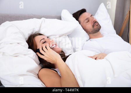 Lächelnde junge Frau spricht auf dem Smartphone neben ihrem Mann schlafend auf dem Bett im Schlafzimmer Stockfoto