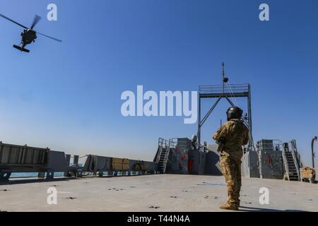 Ein U.S. Army Crew Chief zu C., 2. Bataillon, 211 Allgemeine Unterstützung Aviation Battalion, Minnesota Army National Guard zugeordnet und erwartet die Ankunft eines UH-60L Black Hawk Hubschrauber an Bord des Landing Craft, Utility 2027, USAV Mechanicsville, in den Persischen Golf in der Nähe von der Kuwait Naval Base, Feb 20, 2019. C. in der Firma durchgeführt Aeromedical evacuation Hebezeuge von LCU 2027 Wetter Deck Crew Mitglieder mit einem Hebezeug das Verfahren über eine verankert und bewegten Schiffes vertraut zu machen. Stockfoto