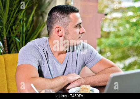 Jungen Kerl Freelancer, der an einem Notebook arbeitet in einem Café. o Der Mann sieht nachdenklich aus dem Fenster Stockfoto