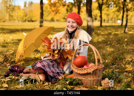 Junge Mädchen in Red Hat glücklich lächelnde. teenager sitzen auf dem Gras, Blätter im Herbst in seinen Händen. In der Nähe von Korb mit Kürbis, Äpfel, gelbe umbre Stockfoto