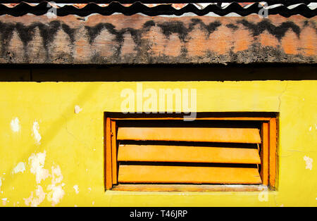 Orange farbigen Fensterläden in einem hellen Gelb gestrichenen Wand mit dunklen Schatten vom Dach aus Holz mit Wellblech Stockfoto