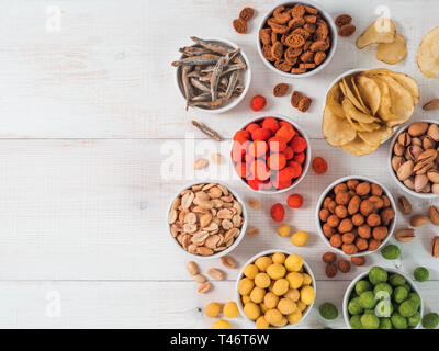 Sortiment von verschiedenen Snack für Bier, Wein, Party. Erdnüsse in Coconut Glasur, grüne vasabi, rote scharfe Chili, gelben Käse Glasur, Chips, Pistazie, Cracker, Fisch auf weiße Holztisch. Platz kopieren Stockfoto