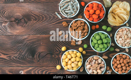 Sortiment von verschiedenen Snack für Bier, Wein, Party. Erdnüsse in Coconut Glasur, grüne vasabi, rote scharfe Chili, gelben Käse Glasur, Chips, Pistazie, Cracker, Fisch auf braune Holztisch. Platz kopieren Stockfoto