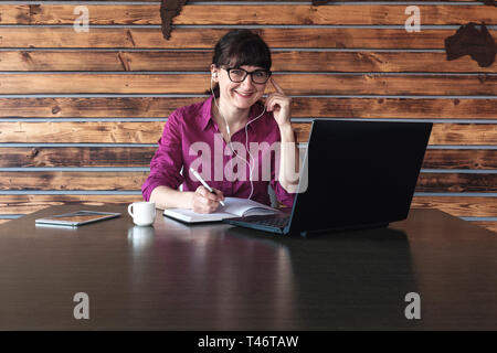 Lächelnd geschäftsfrau Musik hören wie sie arbeitet sich Notizen in einer Zeitschrift beim Lesen aus ihren Laptop Stockfoto