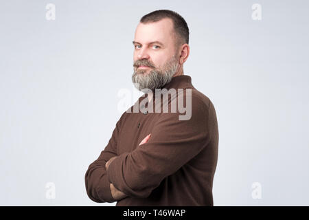 Zuversichtlich älterer Mann mit Bart in Braun t-shirt Kreuzung Hände auf der Brust und Kamera Stockfoto