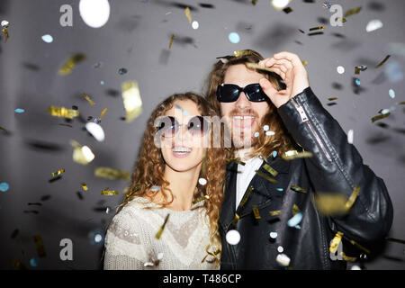Fröhliches, aufgeregtes, hüftjunges Paar mit lockigem Haar, die eine Sonnenbrille trägt, die unter goldenem Konfetti steht und Spaß auf der Party hat Stockfoto