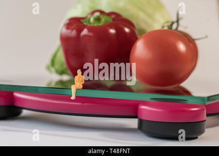 Miniatur Frau Abbildung verlegen auf die digitale elektronische Personenwaage, die für das Gewicht des menschlichen Körpers. Frisches Gemüse bei geringer Tiefenschärfe Hintergrund Stockfoto