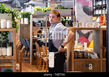 Russland Novosibirsk 2018-11-26 Eine junge männliche Kellner in Uniform, Hemd und schwarze Schürze steht im Restaurant und beobachten die Verkaufsfläche und Kund. Stockfoto