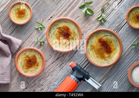 Die Crème Brûlée. Flach auf rustikalen Holztisch mit dieser traditionellen Pudding Dessert in Keramik braun Gerichte und Fackel für den Brennvorgang nicht verwendet Stockfoto