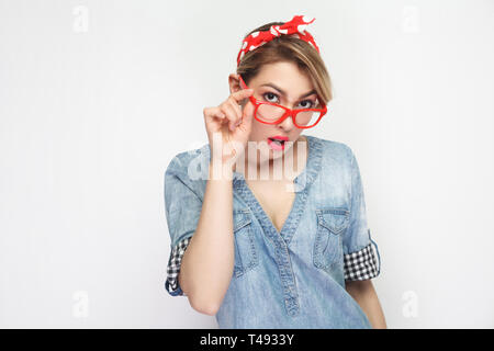 Portrait von aufmerksamen schöne junge Frau in lässigem Blue Denim Shirt mit Make-up und roten Stirnband ständigen Holding Brille und Kamera. Stockfoto