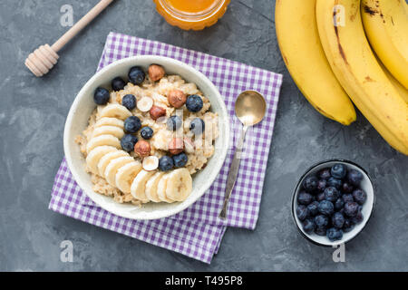 Haferflocken porridge Bowl mit Bananenscheiben, Blaubeeren, Honig und Haselnüssen auf konkreten Hintergrund, Ansicht von oben. Vegane oder vegetarische Frühstück essen, Wiegen Stockfoto