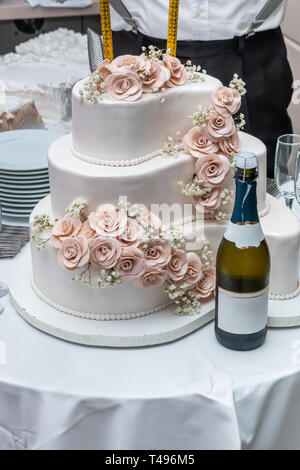 Dreistufige weisse Hochzeitstorte mit Rosen und Champagner auf Tisch im Restaurant eingerichtet. Stockfoto