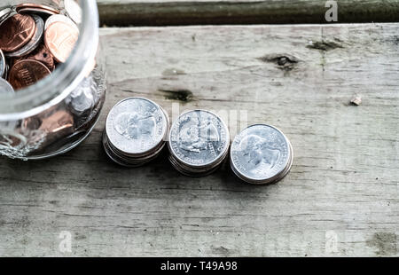 Fokus auf 3 Stapel von Quartalen auf alten Holzboden Board direkt neben einem jar von verschiedenen Münzen. Stockfoto