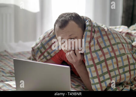 Nachdenklicher Kerl auf dem Bett liegend unter die Abdeckungen und Suchen in den Laptop Stockfoto