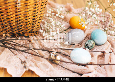 Stilvolle Ostereier mit Blumen und Weidenzweige auf rustikalen Gewebe an Weidenkorb mit Essen im sonnigen Licht. Moderne Ostereier pai Stockfoto