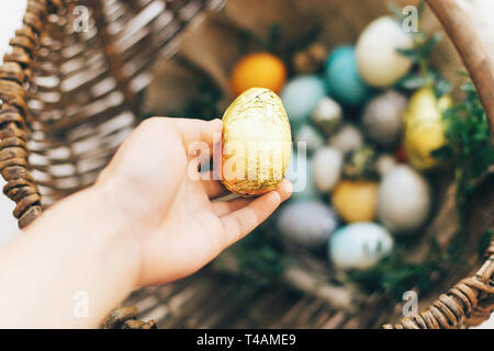 Ostern Jagd Konzept. Hand und goldene Ostereier und stilvolle Eier mit grünen buxus Niederlassungen in rustikalen Weidenkorb auf weißem Holz- Hintergrund. Hap Stockfoto