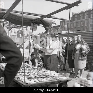 1960, historische, männliche Stall-Inhaber trägt eine Ärmellose Weste sortieren aus seinem Vorrat an Klammern oder Hose hosenträger gurte an seinem Stall, mit den Menschen vor Ort auf, in Stepney, East London, London, England, Großbritannien Stockfoto