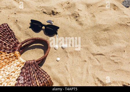 Stilvolle Sonnenbrillen, Stroh Beutel am Sandstrand mit Muscheln, Ansicht von oben mit der Kopie. Sommer Urlaub und Reisen Konzept. Sommer Zubehör und Essent Stockfoto