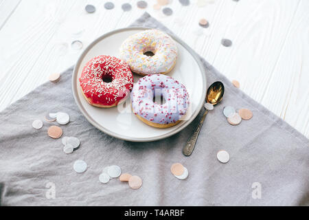 Leckere bunte Donuts mit Streuseln auf stilvolle Platte am weißen Tisch mit Konfetti. Party und Feier Konzept. Keine Diät. Candy Bar bei der Hochzeit r Stockfoto
