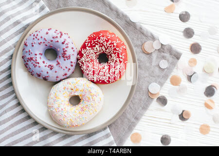 Leckere bunte Donuts mit Streuseln auf stilvolle Platte am weißen Tisch mit Konfetti, flach. Party Konzept. Keine Diät. Candy Bar bei der Hochzeit Rezept Stockfoto