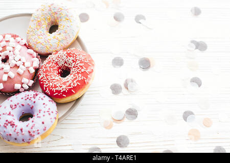 Leckere bunte Donuts mit Streuseln und Marshmallows auf stilvolle Platte am weißen Tisch mit Konfetti. Party Konzept. Keine Diät. Candy Bar bei der Hochzeit Stockfoto