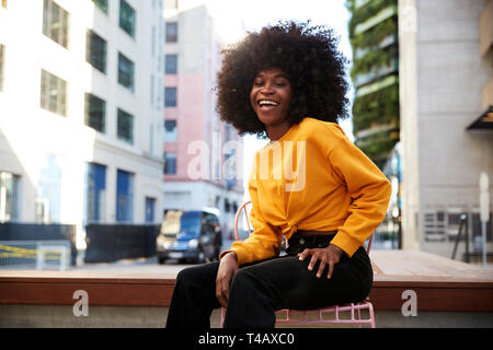 Junge schwarze Frau mit afro Haar sitzt auf einem Stuhl in der Straße an der Kamera lachend Stockfoto