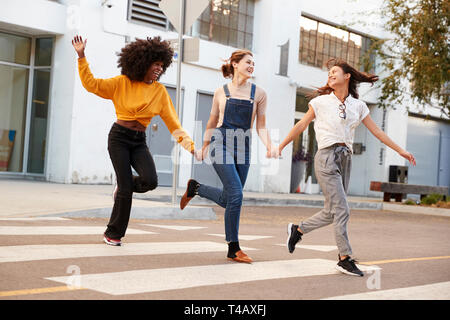 Drei tausendjährigen hip Freundinnen halten sich an den Händen und lachte, als sie über eine Fußgängerbrücke überqueren ausführen Stockfoto