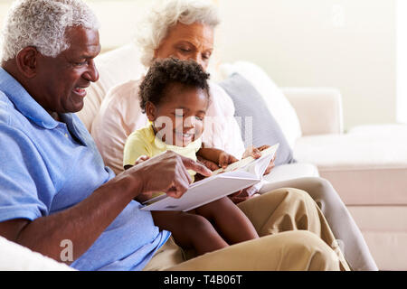 Großeltern Sitzen auf einem Sofa zu Hause mit Baby Enkelin Buch zusammen Lesen Stockfoto