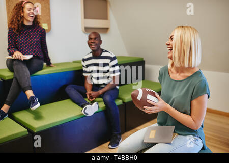 Lachende junge Frau mit einem Fußball beim Sitzen mit Kollegen bei einem zwanglosen Treffen in einem modernen Büro Stockfoto