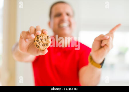 Mittleres Alter Mann essen Chocolate Chips cookies zu Hause sehr zufrieden zeigt mit Finger- und an der Seite Stockfoto