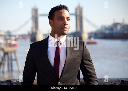 Tausendjährige schwarzer Geschäftsmann mit schwarzen Jacke, weißes Hemd und Krawatte an der Themse Riverside in London, Tower Bridge im Hintergrund, im Vordergrund Schwerpunkt Stockfoto