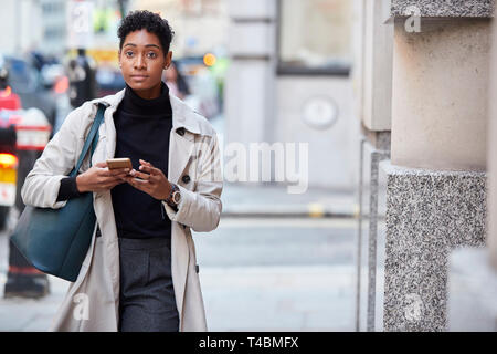 Junge schwarze Geschäftsfrau zu Fuß auf der Straße in London können Sie über ihr Smartphone, Vorderansicht Stockfoto