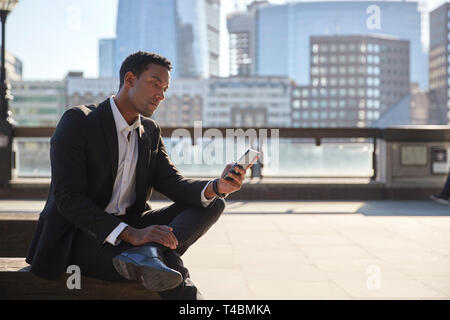 Tausendjährige Geschäftsmann mit schwarzen Anzug und weißes Hemd sitzen auf der Themse Ufer mit Smartphone, in der Nähe Stockfoto