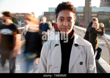Junge schwarze Frau, die über die Millennium Bridge, London, in die Kamera schauen, während Fußgänger durch Bewegungsunschärfe Pass Stockfoto