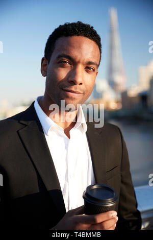 Junge schwarze Unternehmer in Smart Casual Kleidung stehen durch die Themse in London mit einem Takeaway Kaffee, Blick in die Kamera lächeln, Nahaufnahme Stockfoto