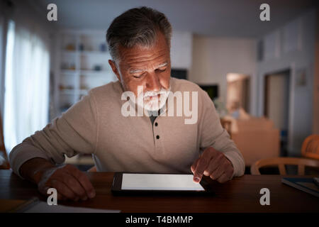 Senior Hispanic Mann an einem Tisch sitzen mit einem Tablet Computer zu Hause am Abend, in der Nähe Stockfoto