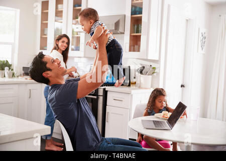 Junger spanischer Familie in der Küche, Papa anheben Baby in der Luft, Mama Kochen am Herd, in der Nähe Stockfoto