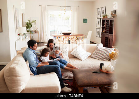 Junger spanischer Familie sitzt auf dem Sofa, ein Buch lesen gemeinsam im Wohnzimmer, vom Eingang gesehen Stockfoto